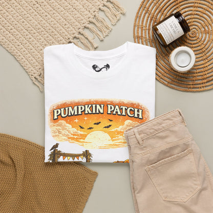 A folded white graphic T-shirt is arranged in a styled flat lay with beige pants, a woven mat, a candle jar, and a small cup on a light neutral background. The shirt graphic reads “PUMPKIN PATCH” over an orange sunset scene with bats and a raven.
