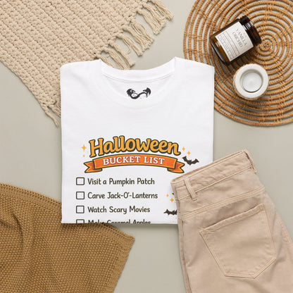 A folded white graphic T-shirt is arranged in a styled flat lay with beige pants, a woven placemat, a small candle, and a jar on a neutral tabletop. The shirt graphic reads 'Halloween Bucket List' with checklist items including visiting a pumpkin patch, carving jack-o'-lanterns, watching scary movies, and making caramel apples.
