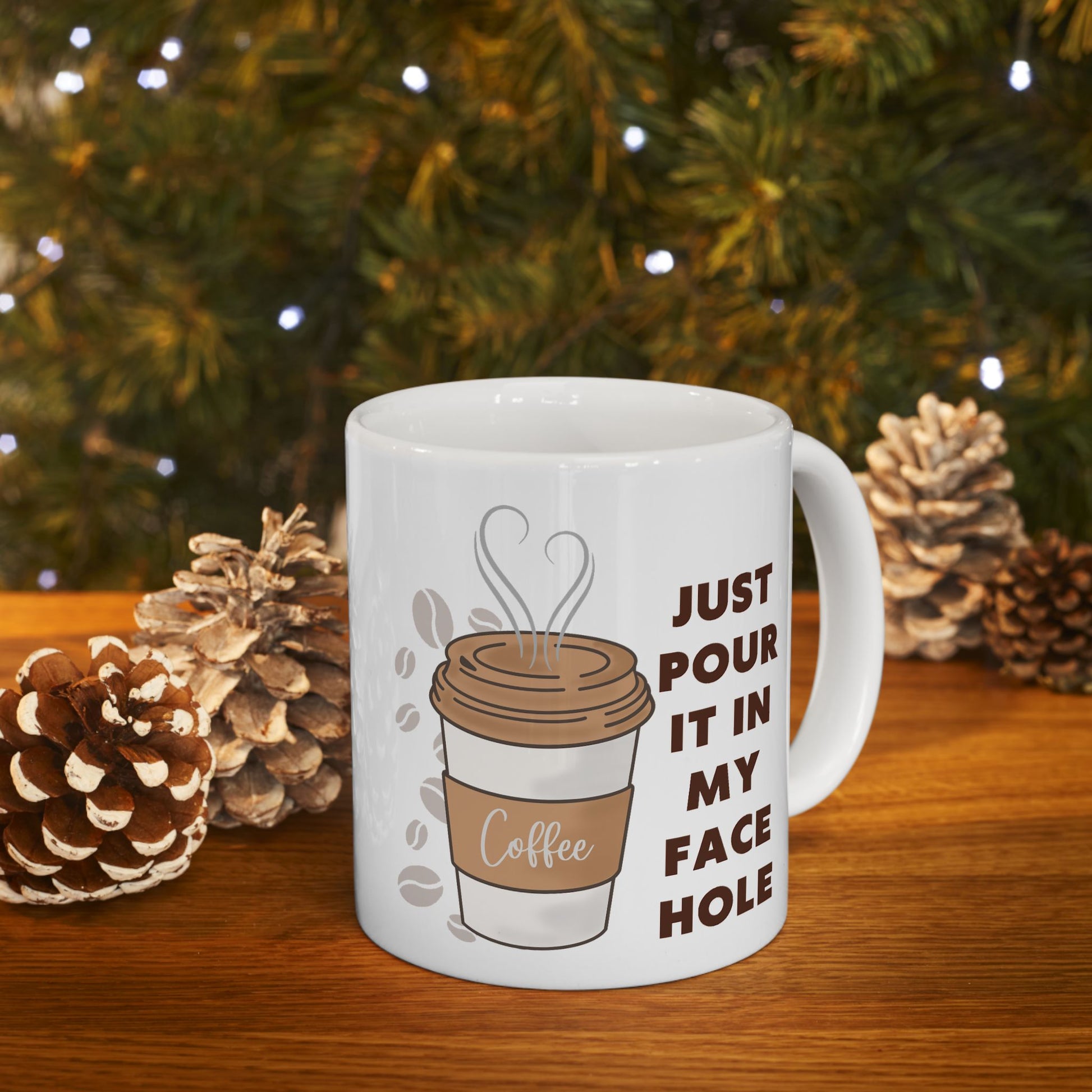 White mug with coffee-themed design and text, placed on a wooden surface with pinecones and a Christmas tree in the background.