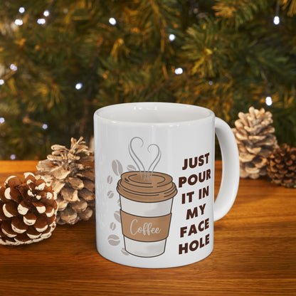 White mug with coffee-themed design and text, placed on a wooden surface with pinecones and a Christmas tree in the background.