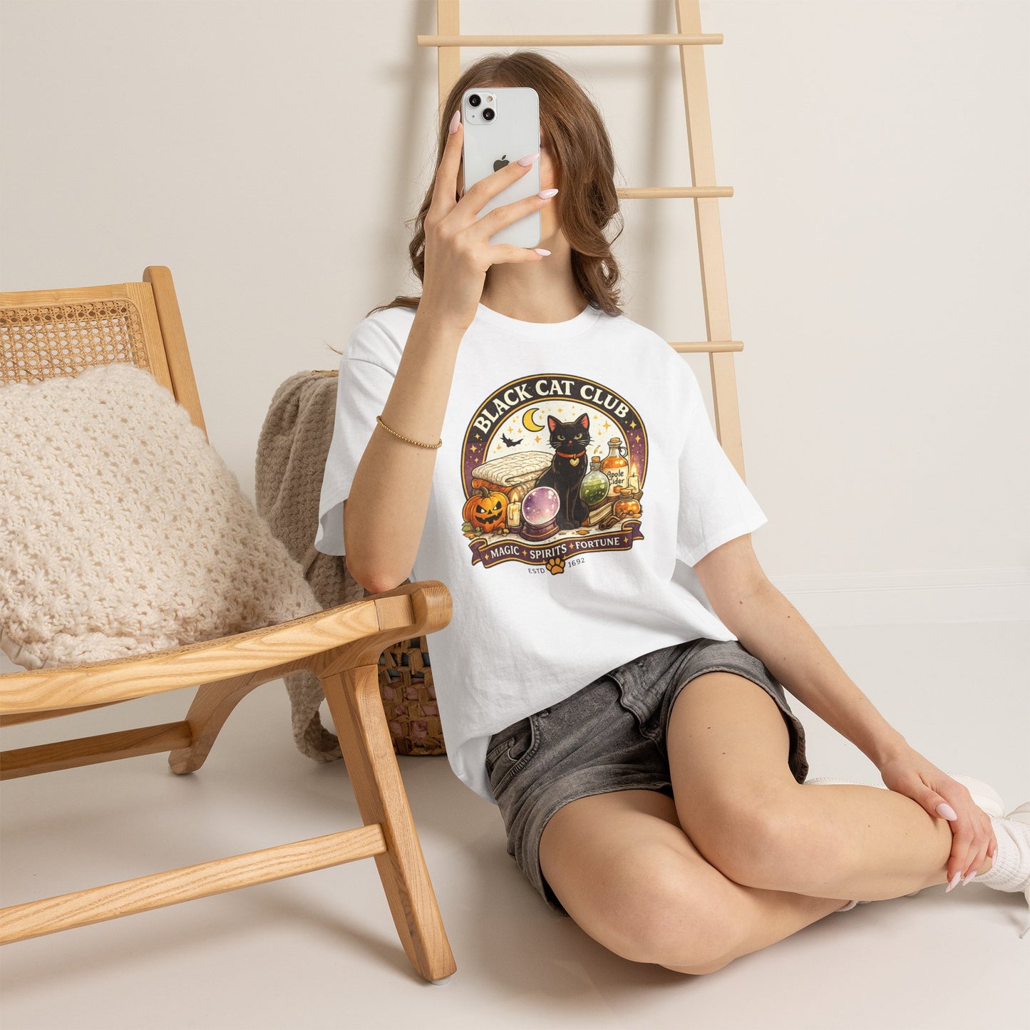 A woman takes a mirror selfie while wearing a white graphic t-shirt with a 'Black Cat Club' design featuring a black cat, a pumpkin, and a witch, paired with denim shorts in a cozy room setting.