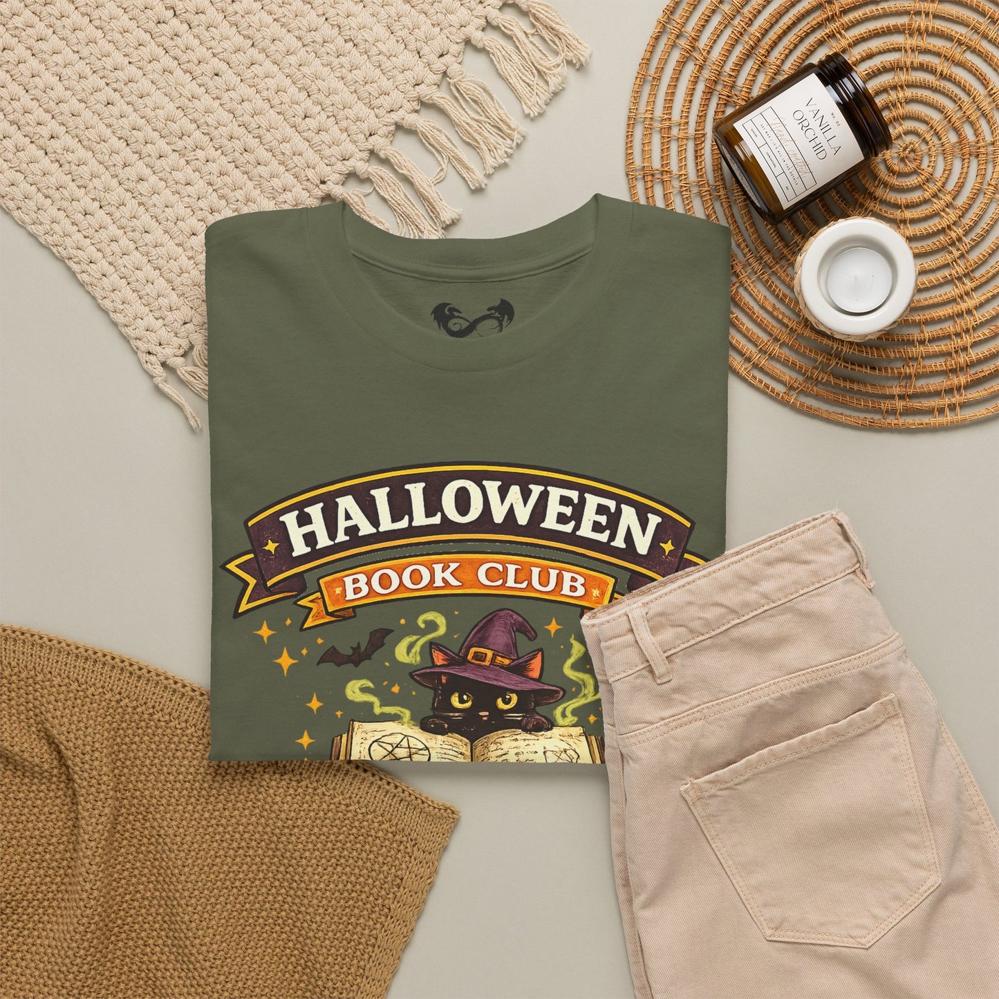 An olive green graphic t-shirt featuring a 'Halloween Book Club' design with a black cat in a witch hat, styled in a cozy flat-lay with beige pants, a knit blanket, a candle, and a jar on a neutral background.