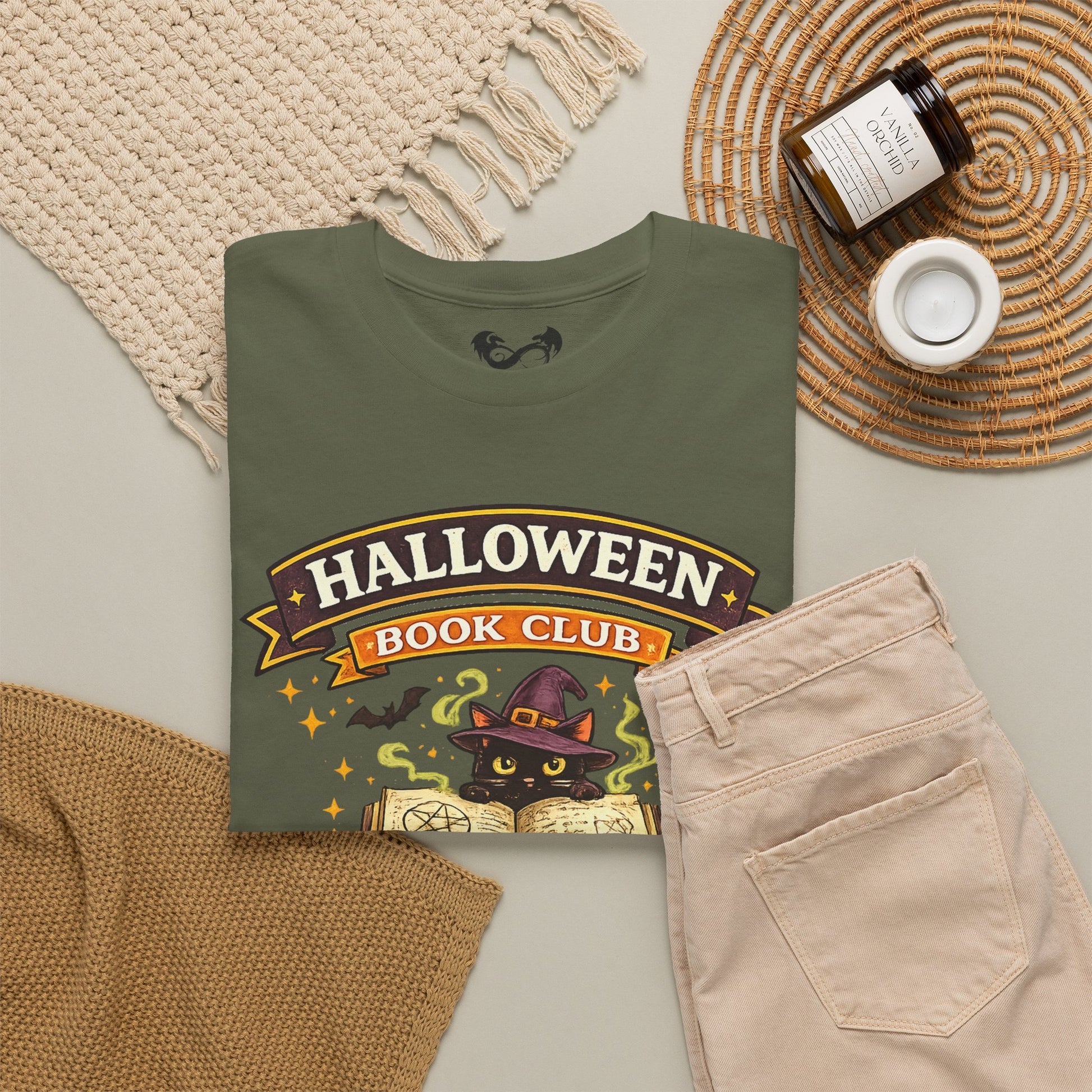 An olive green graphic t-shirt featuring a 'Halloween Book Club' design with a black cat in a witch hat, styled in a cozy flat-lay with beige pants, a knit blanket, a candle, and a jar on a neutral background.