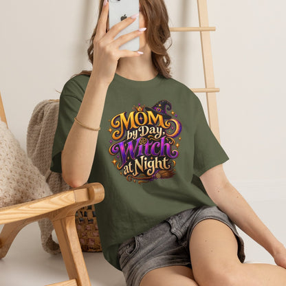 A woman takes a mirror selfie while wearing an olive green graphic t-shirt that reads 'MOM my Day Witch at Night' in a colorful, stylized font. She is sitting on a light-colored floor next to a wooden chair with a knit blanket, with a wooden ladder leaning against the wall in the background.