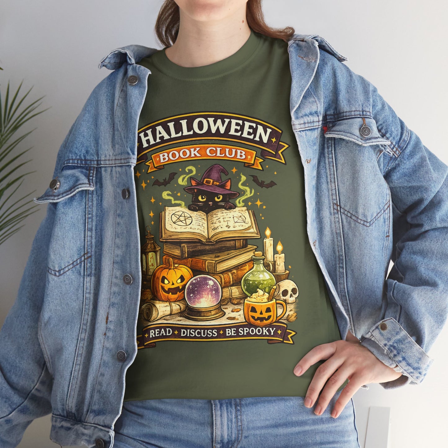 A person wearing an olive green graphic T-shirt under an open light-wash denim jacket. The shirt features a Halloween-themed book club illustration with a black cat, witch hat, books, pumpkins, candles, a crystal ball, and text reading 'HALLOWEEN BOOK CLUB' and 'READ • DISCUSS • BE SPOOKY'.