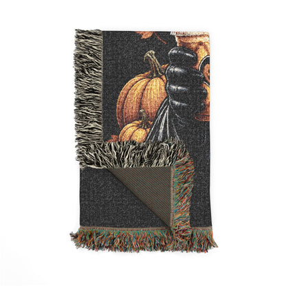 A dark charcoal woven throw blanket with fringe, featuring a printed autumn harvest design with pumpkins and a hand, shown on a white background.