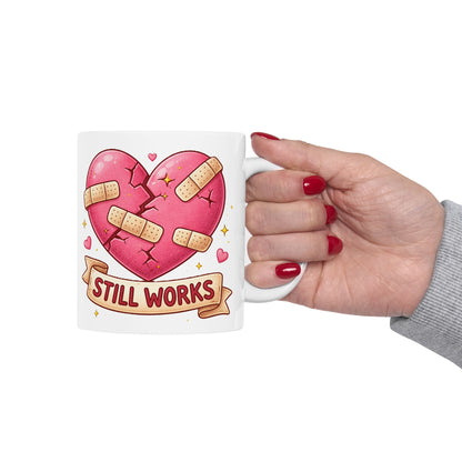 Mug with a heart design and 'STILL WORKS' text held by a hand with red nail polish.