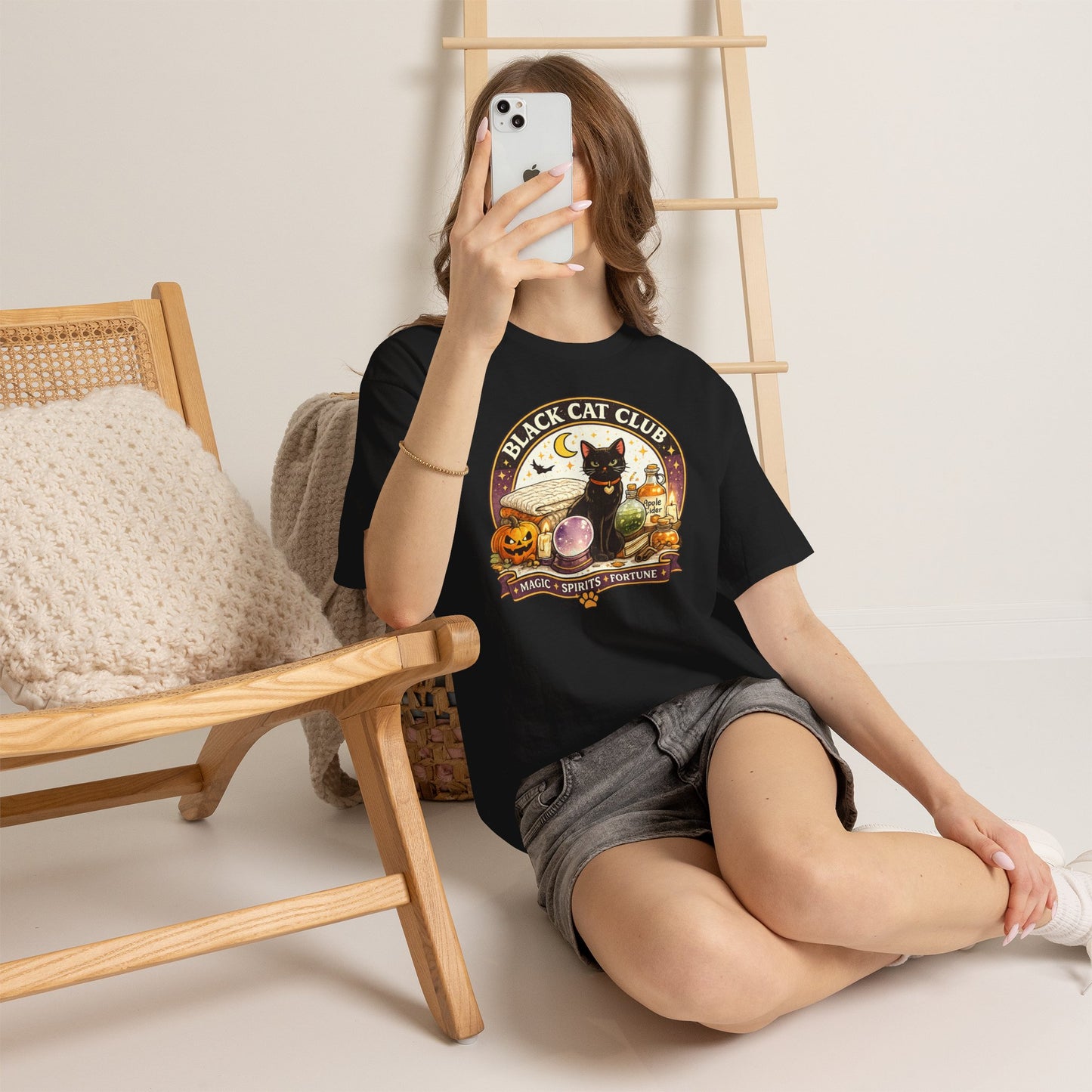 A woman sitting on the floor takes a mirror selfie while wearing a black graphic t-shirt with a 'Black Cat Club' design. The setting is a cozy room with a wooden chair, a knit pillow, and a wooden ladder against a light beige wall.