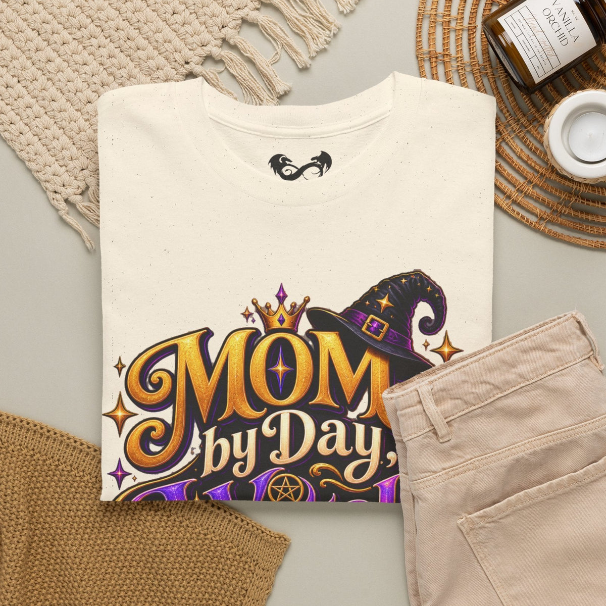 A folded cream-colored t-shirt with a 'MOM by Day' witch graphic, styled in a flat lay with beige pants, a knit blanket, a candle, and a small cup on a neutral background.