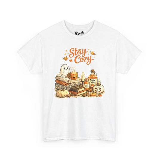 A white short-sleeve crewneck t-shirt featuring a colorful graphic print on the front. The graphic shows a ghost sitting on a stack of books next to pumpkins, a potion bottle, and candles under the text 'Stay Cozy' in orange.