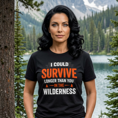 A woman stands outdoors in a forested mountain setting wearing a black graphic T-shirt with the slogan 'I COULD SURVIVE LONGER THAN YOU IN THE WILDERNESS' in white and orange text, paired with blue jeans.