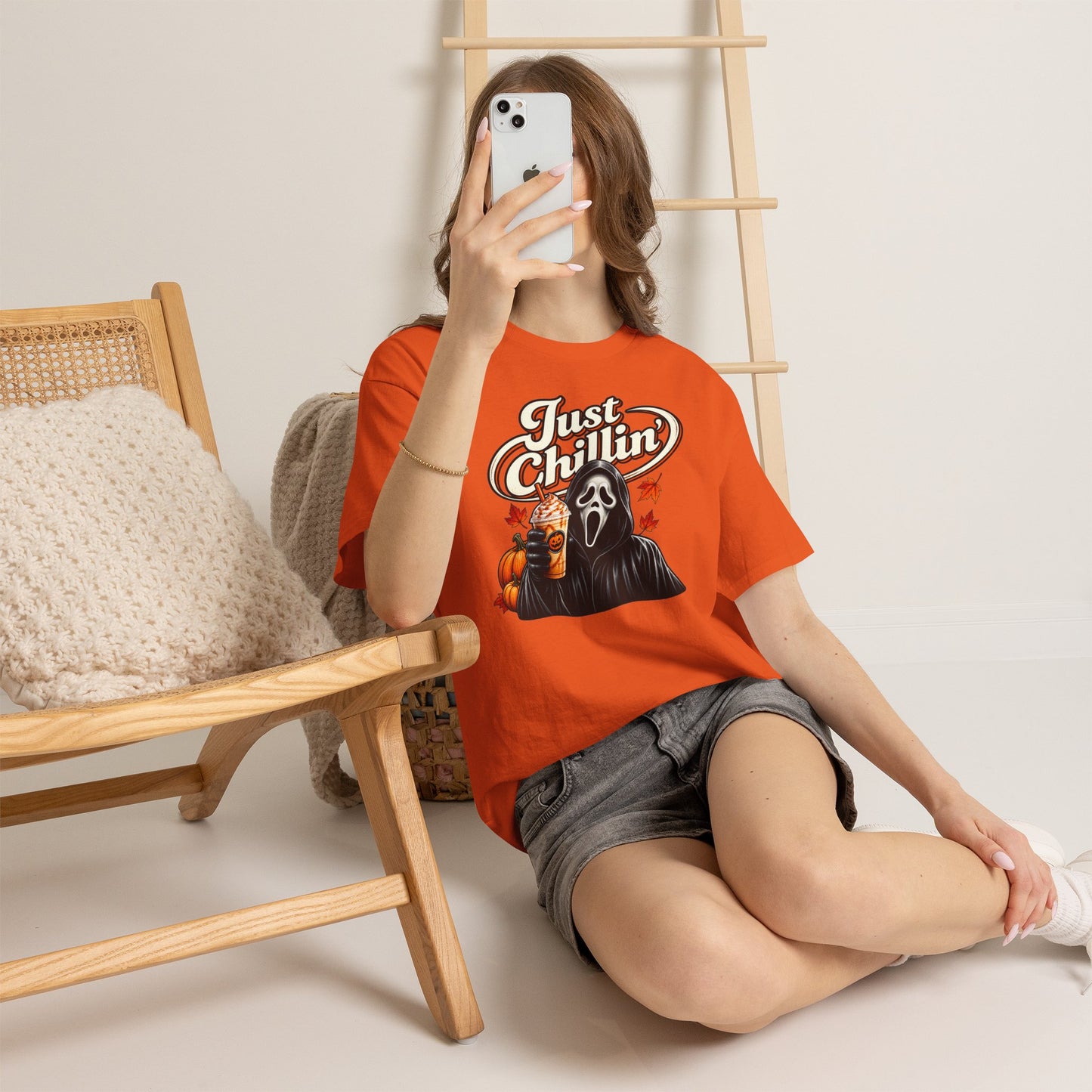 A woman takes a mirror selfie while wearing a bright orange graphic t-shirt featuring the Ghostface character and the text 'Just Chillin''. She is sitting on a light-colored floor next to a wooden chair with a knit pillow.