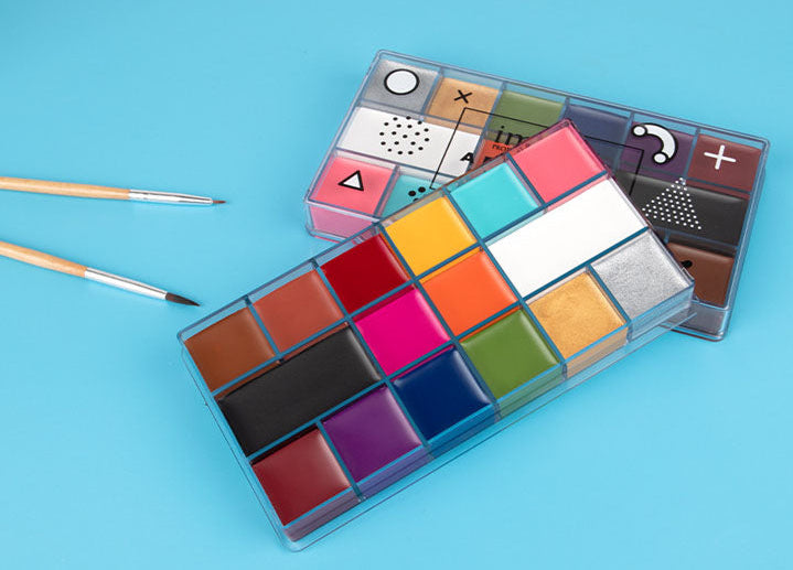 A clear plastic watercolor paint palette with multiple color pans, shown open on a bright blue background, alongside two small paintbrushes and a second closed palette displaying printed geometric color charts.