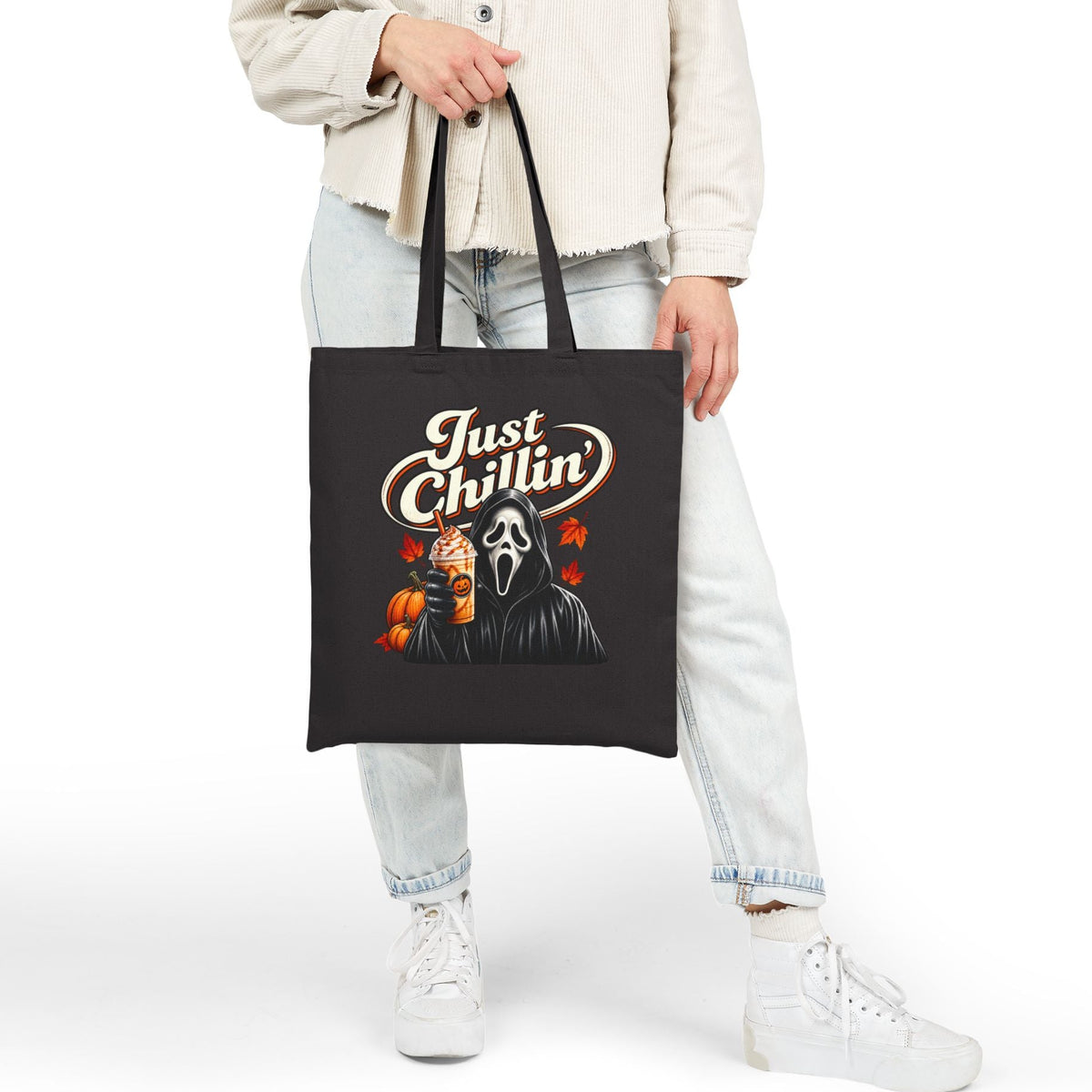 A person wearing a light beige jacket, light wash jeans, and white sneakers holds a black canvas tote bag. The bag features a graphic print with the text 'Just Chillin'' in a stylized cream font, above a depiction of Ghostface holding a pumpkin next to a black figure with red roses.
