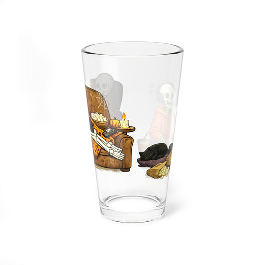 A clear pint glass featuring a Halloween-themed illustration of a skeleton armchair, a black cat, and ghostly figures on a white background.