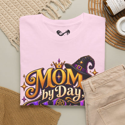 A folded light pink T-shirt with a colorful 'MOM by Day' graphic featuring a witch hat and gold lettering, styled in a flat lay with beige pants, a woven blanket, a candle, and a small cup on a neutral background.