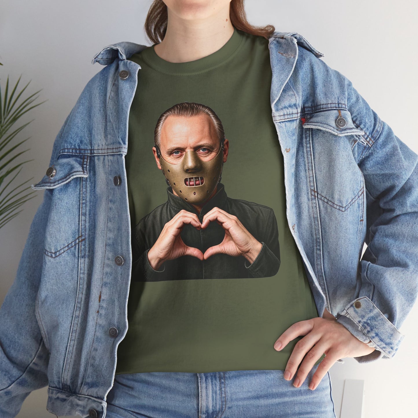 A person wearing an olive green graphic t-shirt with a print of a man in a mask making a heart shape with his hands, styled with an open light-wash denim jacket.