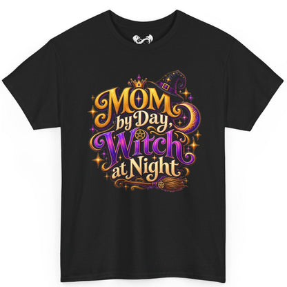 A black short-sleeve crewneck t-shirt featuring a colorful graphic print on the front with the text 'MOM by Day, Witch at Night' in stylized gold, purple, and orange lettering with a witch hat, broom, and stars.
