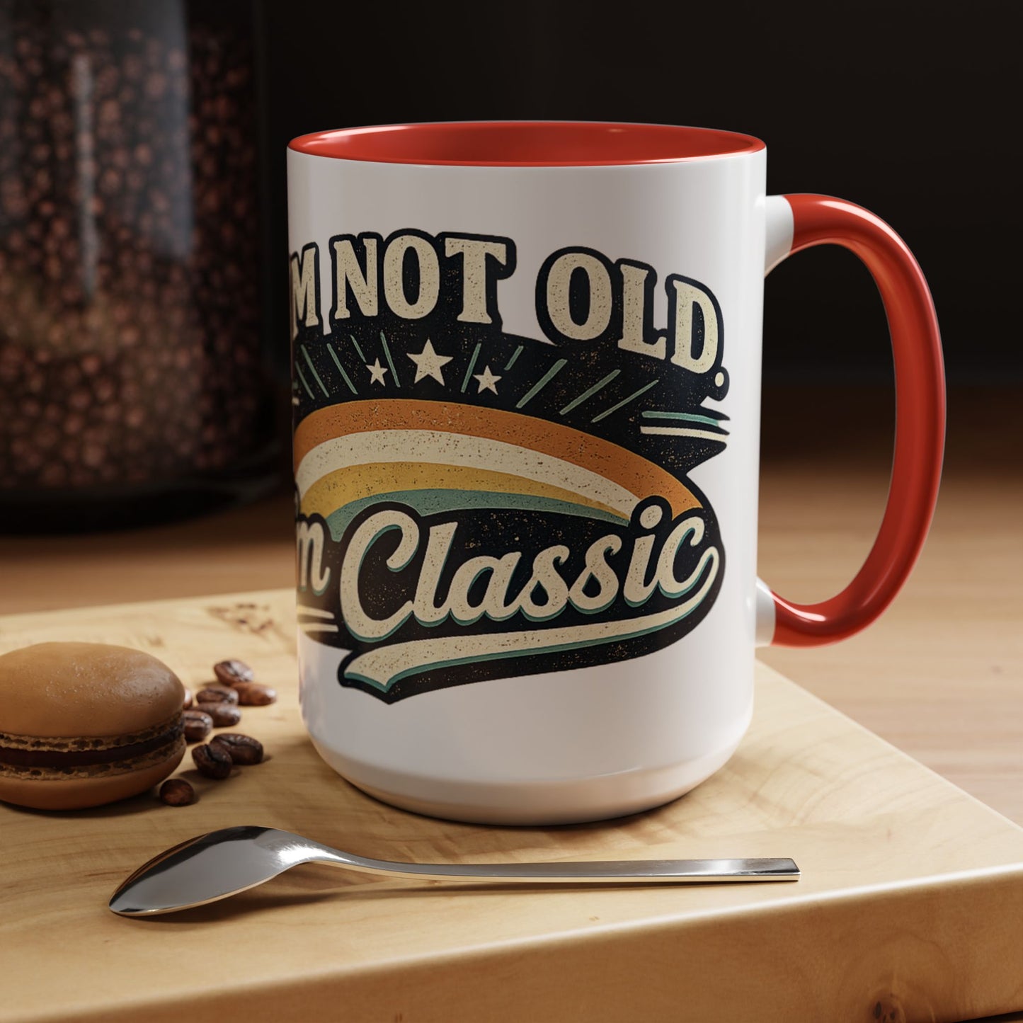 White mug with red interior and handle, featuring 'I'm not old, I'm classic' text, on a wooden surface with coffee beans and a macaron.