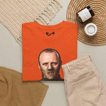 An orange folded T-shirt with a graphic print of Jason Voorhees' face on the front, styled in a flat lay with beige pants, a knit blanket, a candle jar, and a woven placemat on a light gray surface.