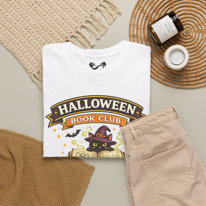 A folded white graphic T-shirt is styled in a flat lay with beige pants, a knit blanket, a woven mat, a candle jar, and a small cup. The shirt graphic reads "HALLOWEEN BOOK CLUB" with a black cat wearing a witch hat and stars.