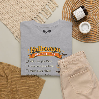 A folded light gray T-shirt with a Halloween bucket list graphic, styled in a flat lay with beige pants, a woven placemat, a small candle, and a jar on a neutral background.
