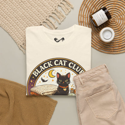 A folded cream graphic T-shirt is styled in a flat lay with beige pants, a woven blanket, a candle jar, and a small white cup on a light gray surface. The shirt graphic reads 'BLACK CAT CLUB' around an illustration of a black cat under a crescent moon.