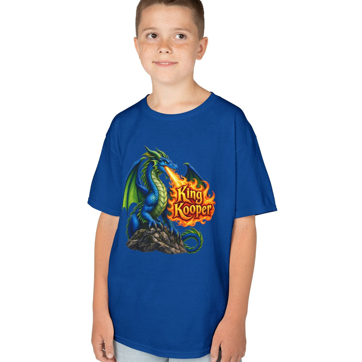 A young boy modeling a royal blue t-shirt with a colorful graphic of a green and blue dragon breathing fire, with the text 'King Kooper' in orange flames. The boy is standing against a solid white background.