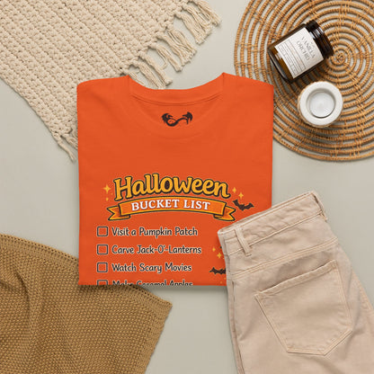 An orange folded T-shirt with a Halloween bucket list graphic, styled in a flat lay with beige pants, a woven placemat, a small candle jar, and a knit textile on a light neutral background.
