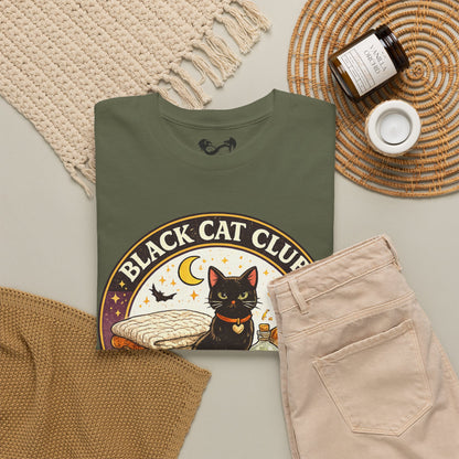 An olive green t-shirt with a 'Black Cat Club' graphic print, styled in a cozy flat-lay arrangement with beige pants, a knit blanket, a candle, and a jar of soap on a neutral background.
