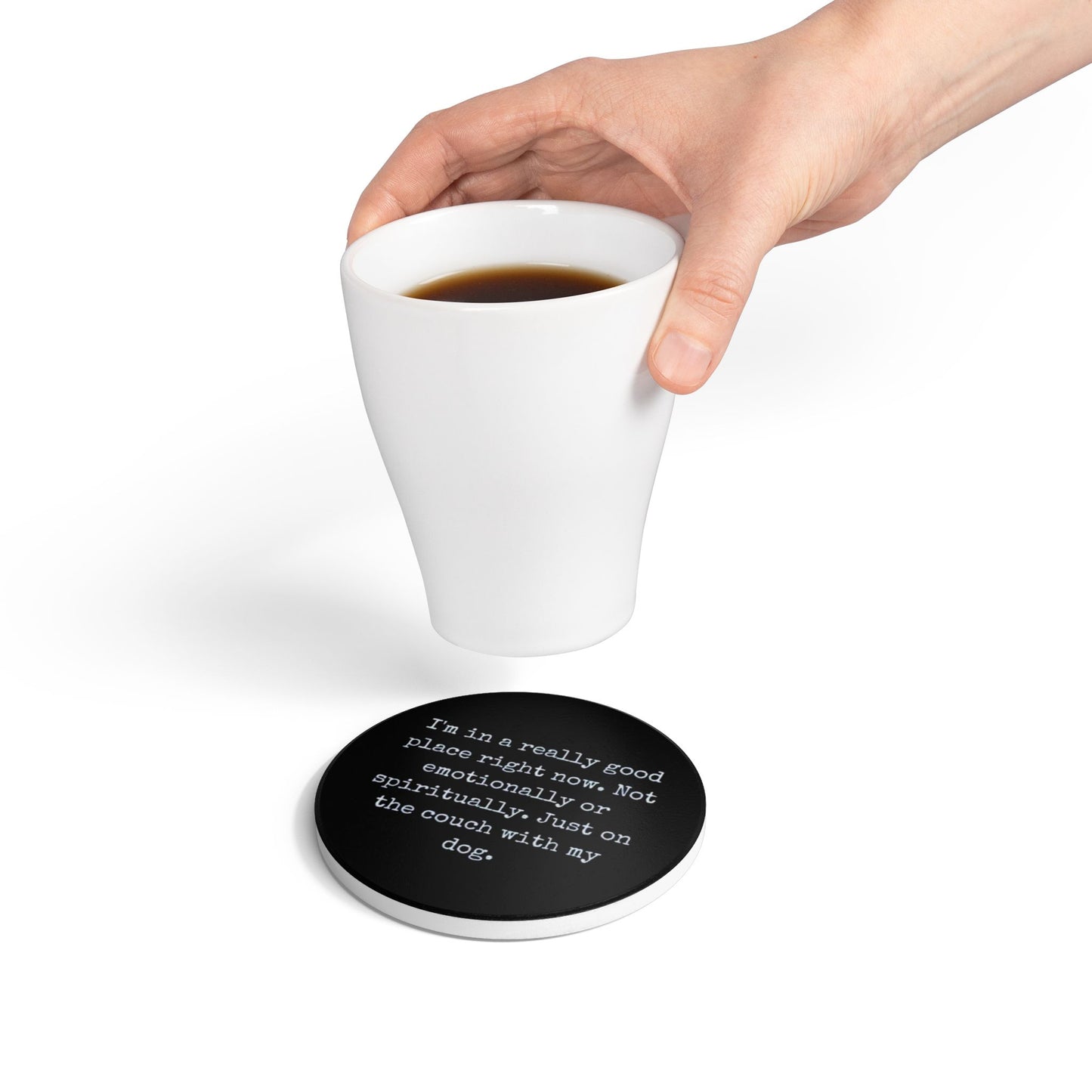 Hand holding a white mug with a black coaster featuring text on a white background