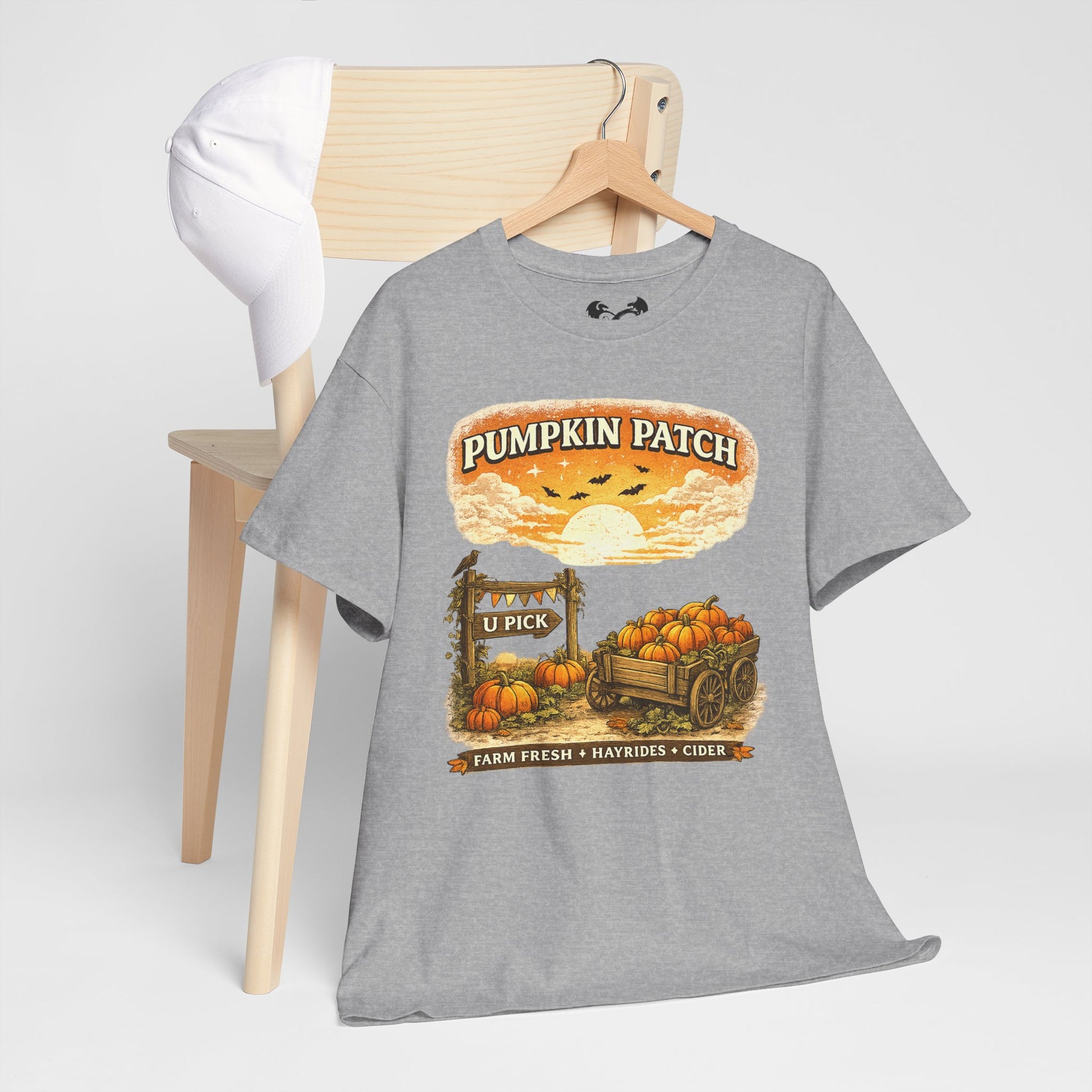 A heather gray graphic t-shirt hanging on a wooden hanger from a light wood chair. The shirt features a fall-themed 'Pumpkin Patch' graphic with pumpkins, a wagon, and a sunset.