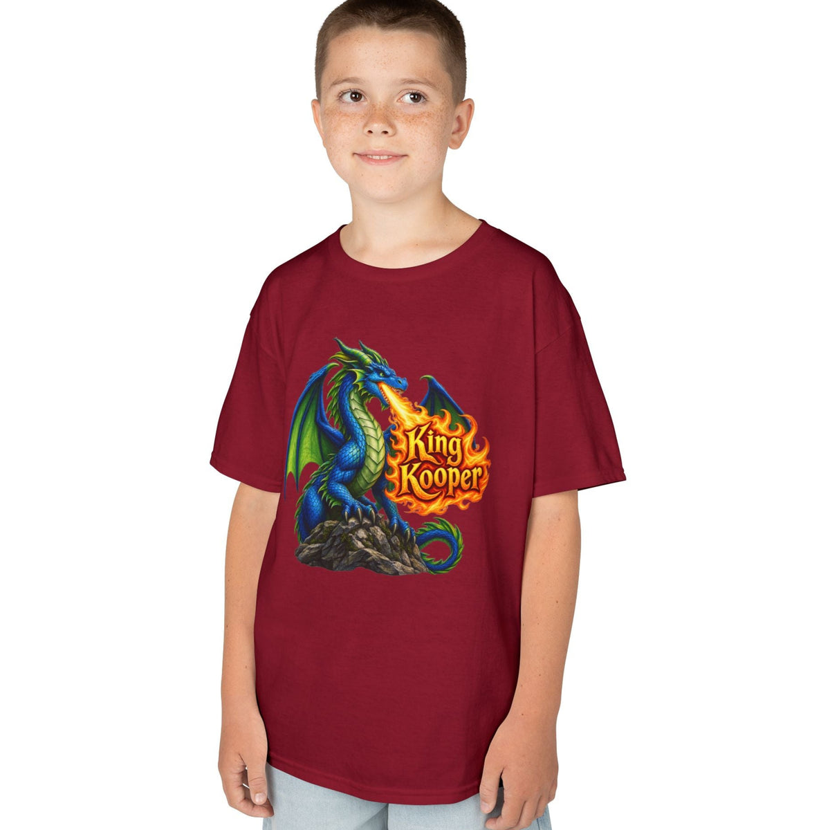 A child wearing a short-sleeve crewneck t-shirt in maroon with a colorful graphic of a blue and green dragon breathing fire, with the text 'King Kooper' in yellow/orange flames. The child is also wearing light blue jeans, set against a plain white background.