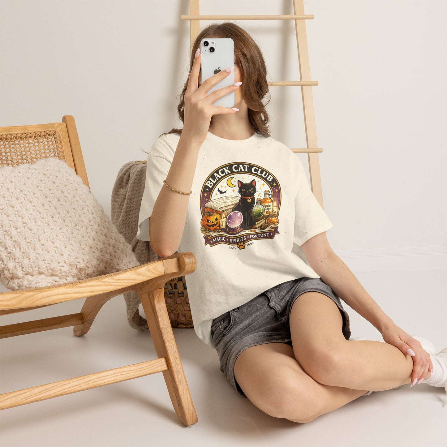 A woman takes a mirror selfie while wearing a cream-colored graphic t-shirt with a 'Black Cat Club' Halloween design and gray denim shorts, sitting on a light-colored floor next to a wooden chair.