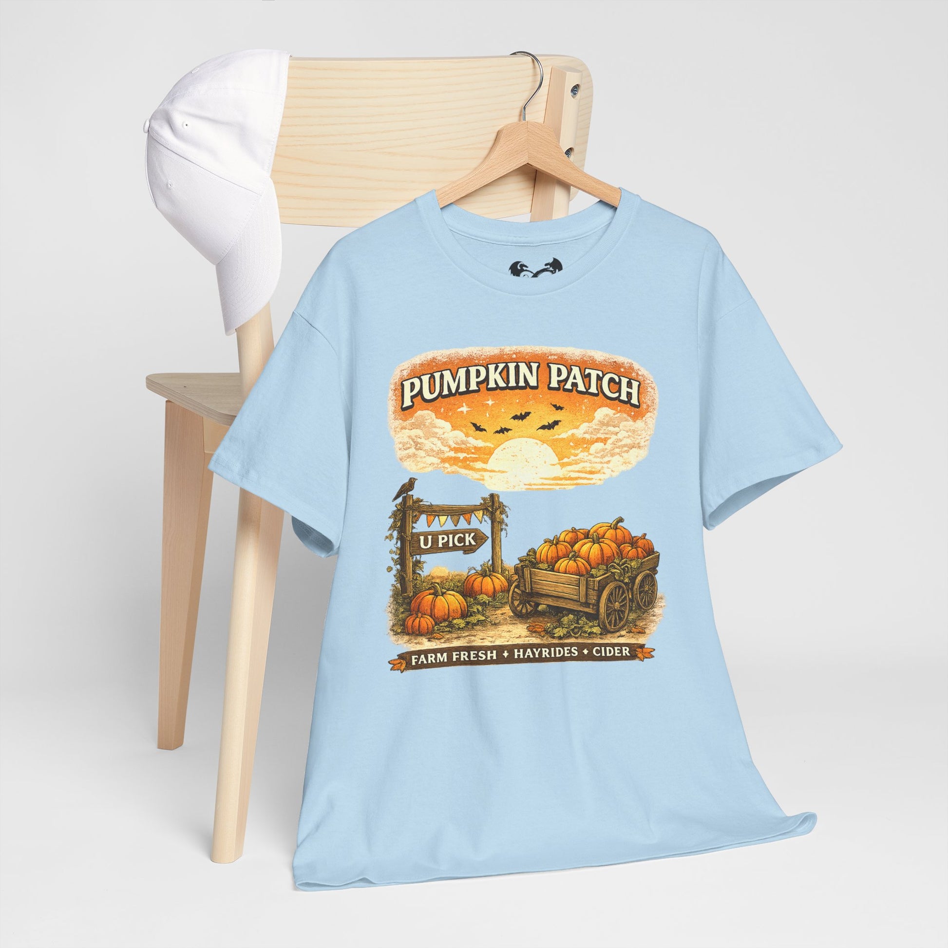 A light blue graphic t-shirt on a wooden hanger, styled on a light wood chair with a white cap in the background. The shirt features a 'Pumpkin Patch' autumn graphic with pumpkins, a wagon, and a sunset.