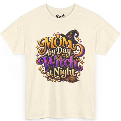 An off-white short-sleeve t-shirt with a colorful graphic on the front that reads 'MOM by Day, Witch at Night' in stylized purple, orange, and gold lettering with a witch hat and broomstick illustration.