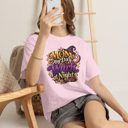 A woman sitting on the floor in a room takes a mirror selfie while wearing a light pink t-shirt with a 'MOM, My Day As A Witch Is Nights' graphic print. She is wearing denim shorts and white sneakers, with a wooden chair and ladder in the background.