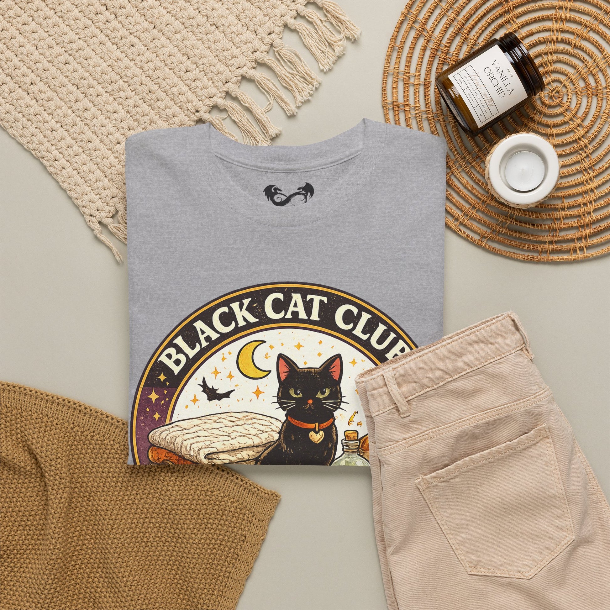 A folded light gray T-shirt with a large circular 'Black Cat Club' graphic featuring a black cat illustration, styled in a flat lay with beige pants, a woven blanket, a candle jar, and a small cup on a neutral background.