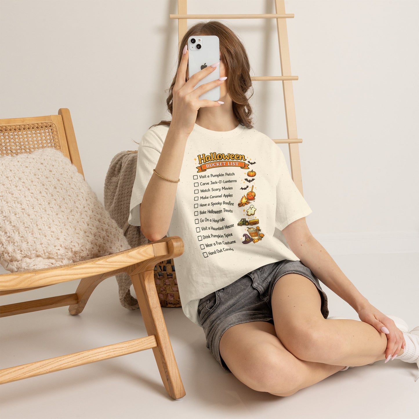 A woman sits on the floor next to a wooden chair, taking a mirror selfie while wearing a cream-colored t-shirt with a 'Halloween Checklist' graphic and gray denim shorts.