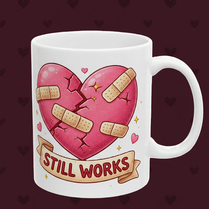 White mug with a pink heart with bandages and 'STILL WORKS' text on a white background