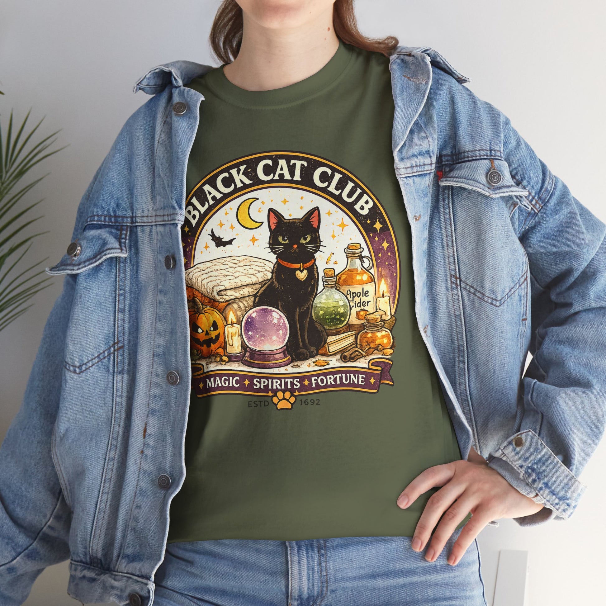 A woman wearing an olive green graphic t-shirt with a 'Black Cat Club' Halloween design, styled with an open light-wash denim jacket.