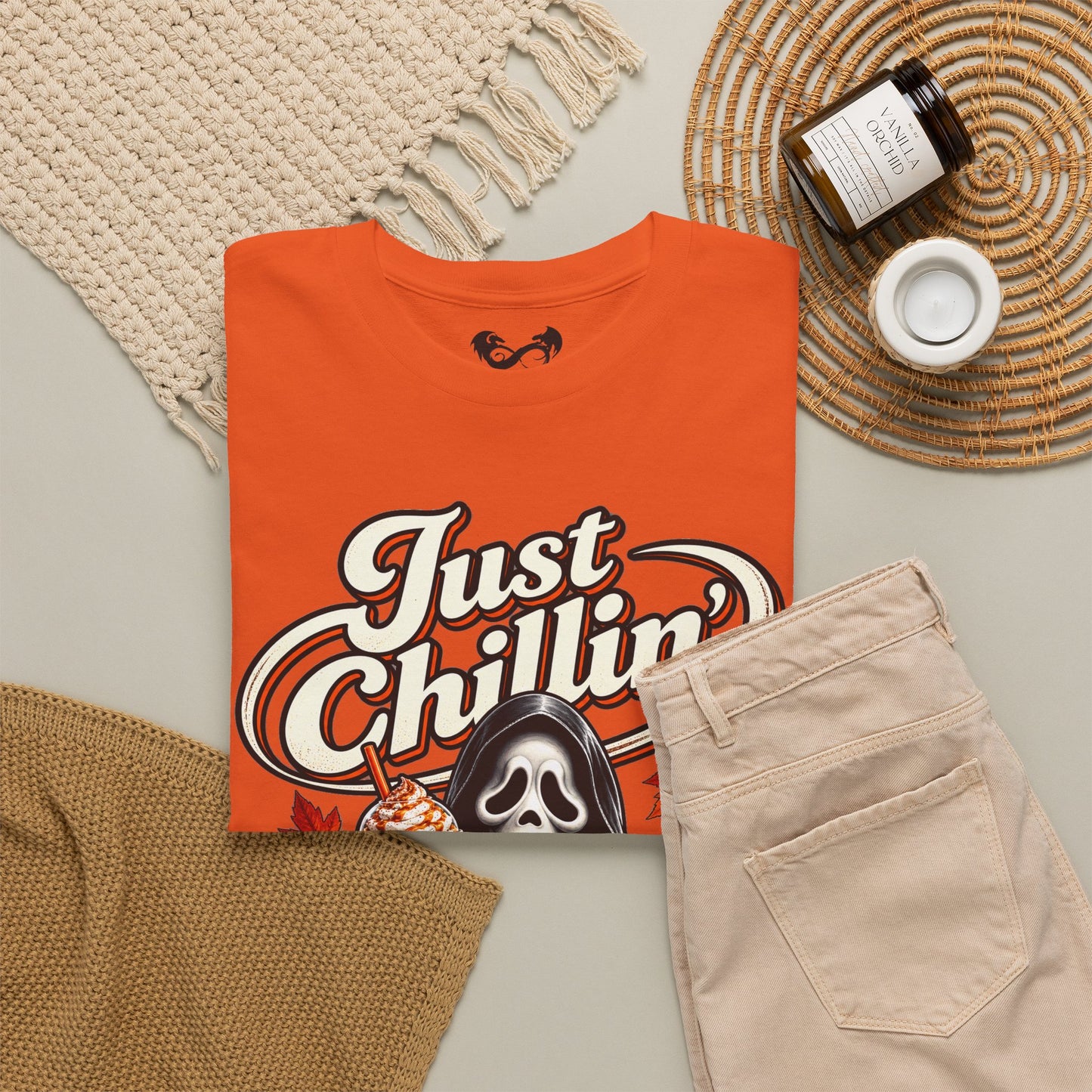 An orange graphic t-shirt with a 'Just Chillin'' Ghostface design, styled in a flat lay with beige pants, a knit blanket, a candle, and a woven placemat on a light gray background.