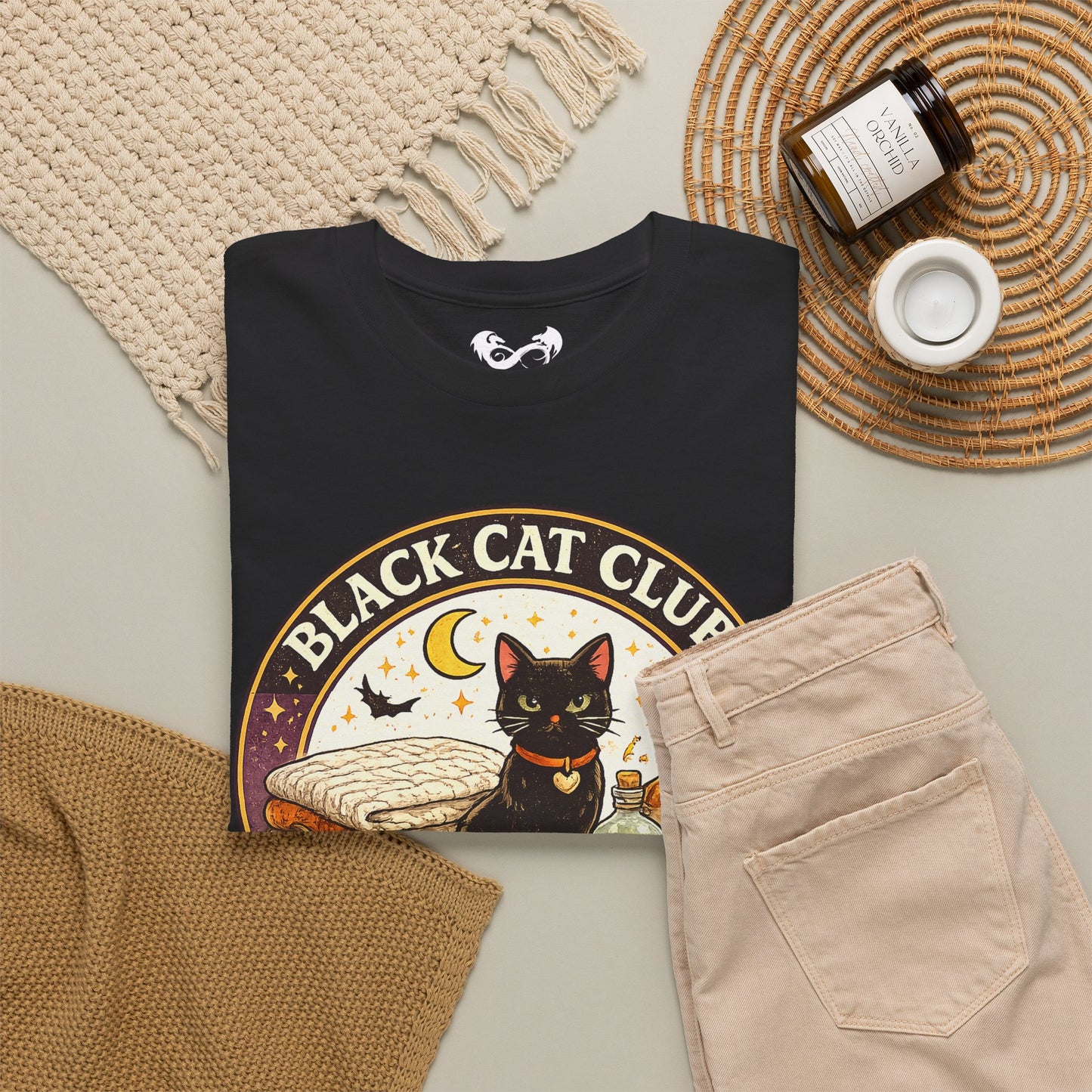 A folded black graphic T-shirt is arranged in a styled flat lay with beige pants, a woven blanket, a candle jar, and a small cup on a light neutral background. The shirt graphic shows a black cat, crescent moon, stars, and the text 'BLACK CAT CLUB'.