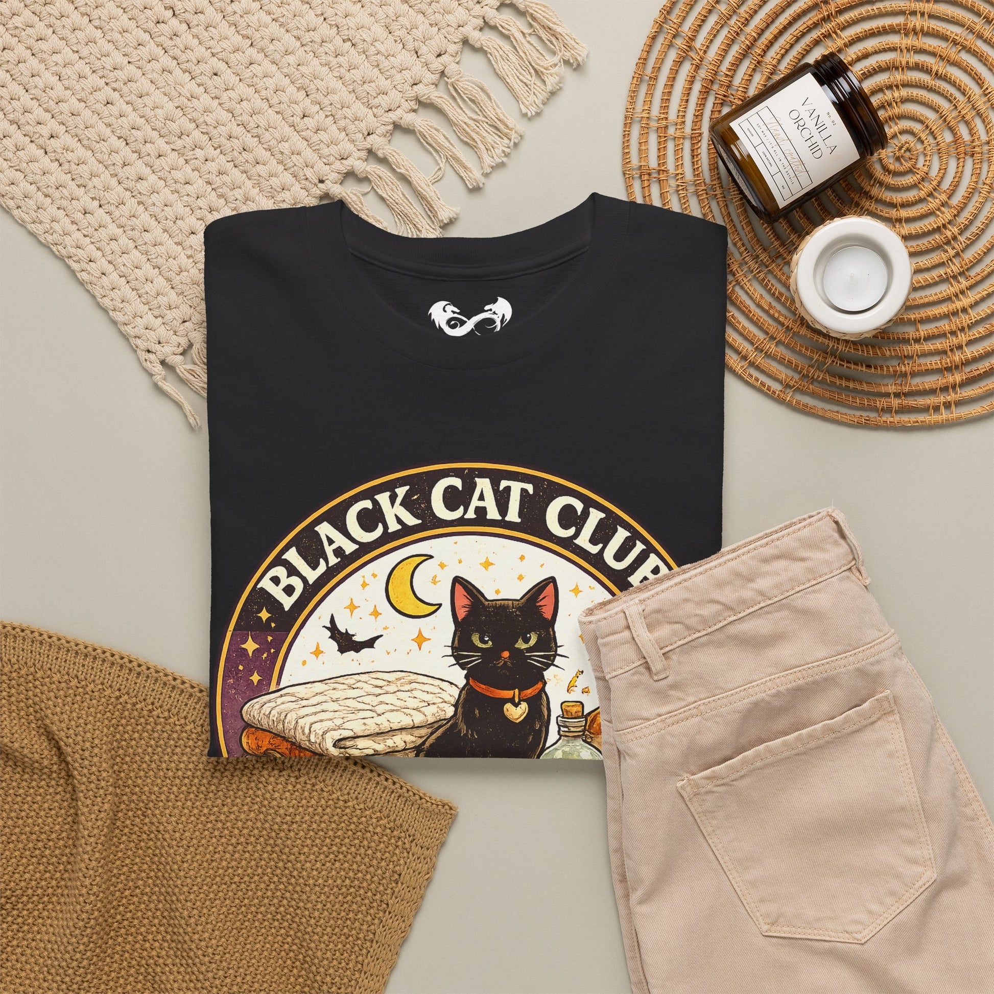 A folded black graphic T-shirt is arranged in a styled flat lay with beige pants, a woven blanket, a candle jar, and a small cup on a light neutral background. The shirt graphic shows a black cat, crescent moon, stars, and the text 'BLACK CAT CLUB'.