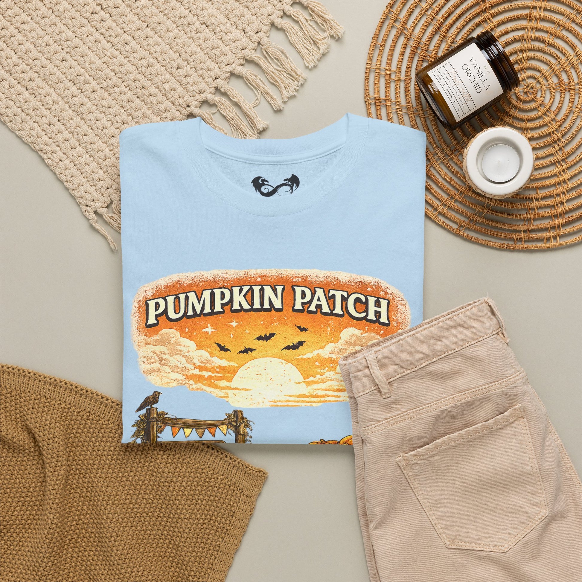 A folded light blue T-shirt with a 'Pumpkin Patch' Halloween graphic, styled in a flat lay with beige pants, a knit blanket, a candle jar, and a woven placemat on a neutral background.