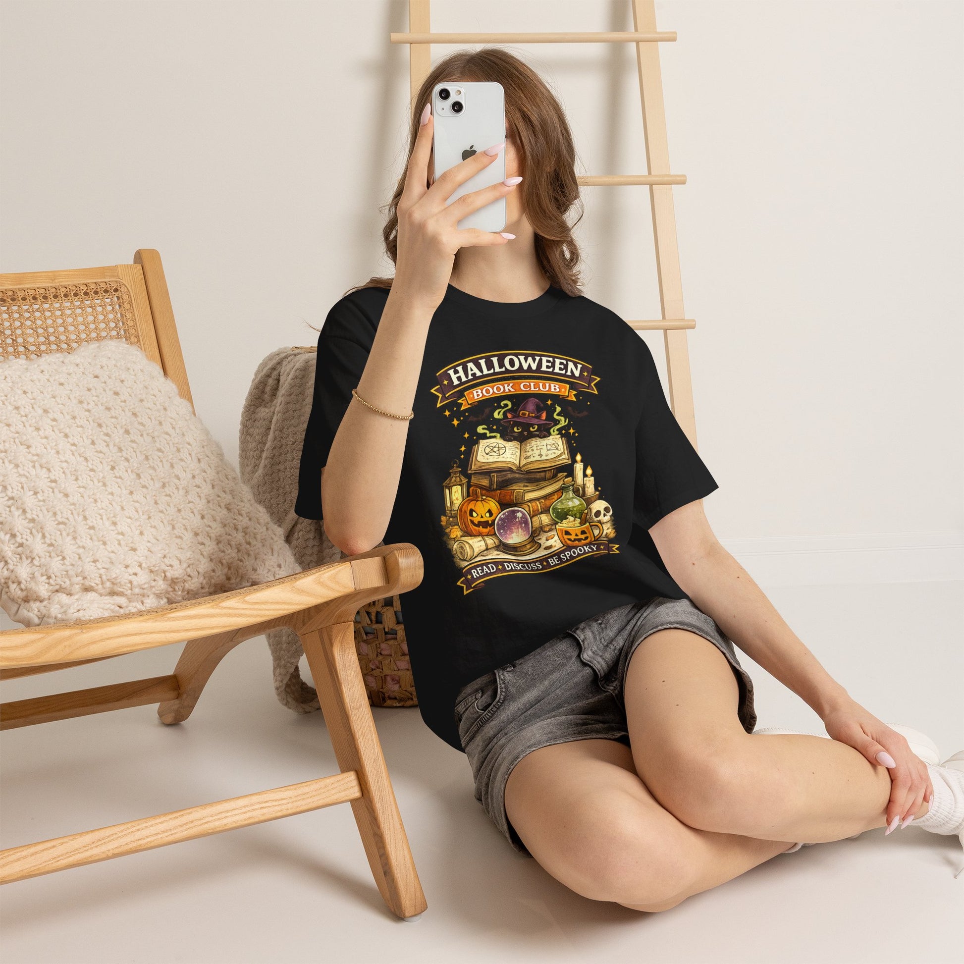 A woman takes a mirror selfie while wearing a black graphic t-shirt with a 'Halloween Book Club' design featuring a stack of books, a skull, and pumpkins. She is sitting on a white floor next to a wooden chair with a knit pillow. Black Cat Halloween Book Club Comfortable Cotton T-Shirt