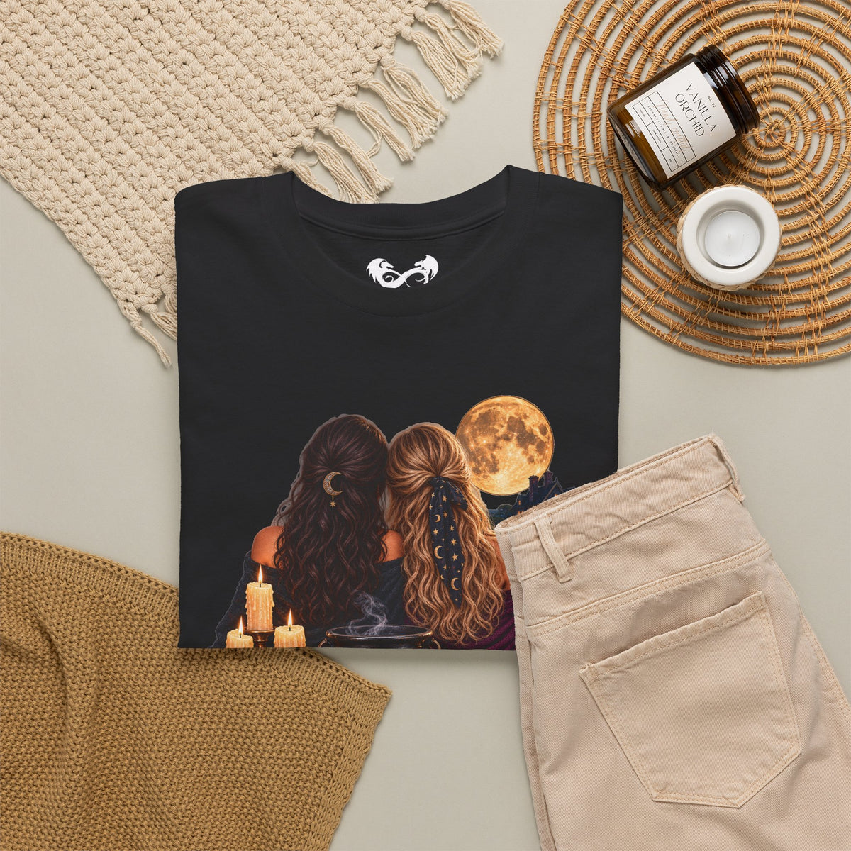 A black graphic t-shirt featuring an illustration of two women with long hair facing a full moon, styled in a cozy flat-lay with beige pants, a knit blanket, a candle, and a jar on a neutral background.