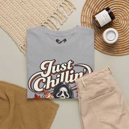 A folded heather gray graphic T-shirt is styled in a flat lay with beige pants, a woven rug, a candle jar, and a small cup on a neutral background. The shirt features a large retro-style graphic reading 'Just Chillin'' with a ghost illustration.