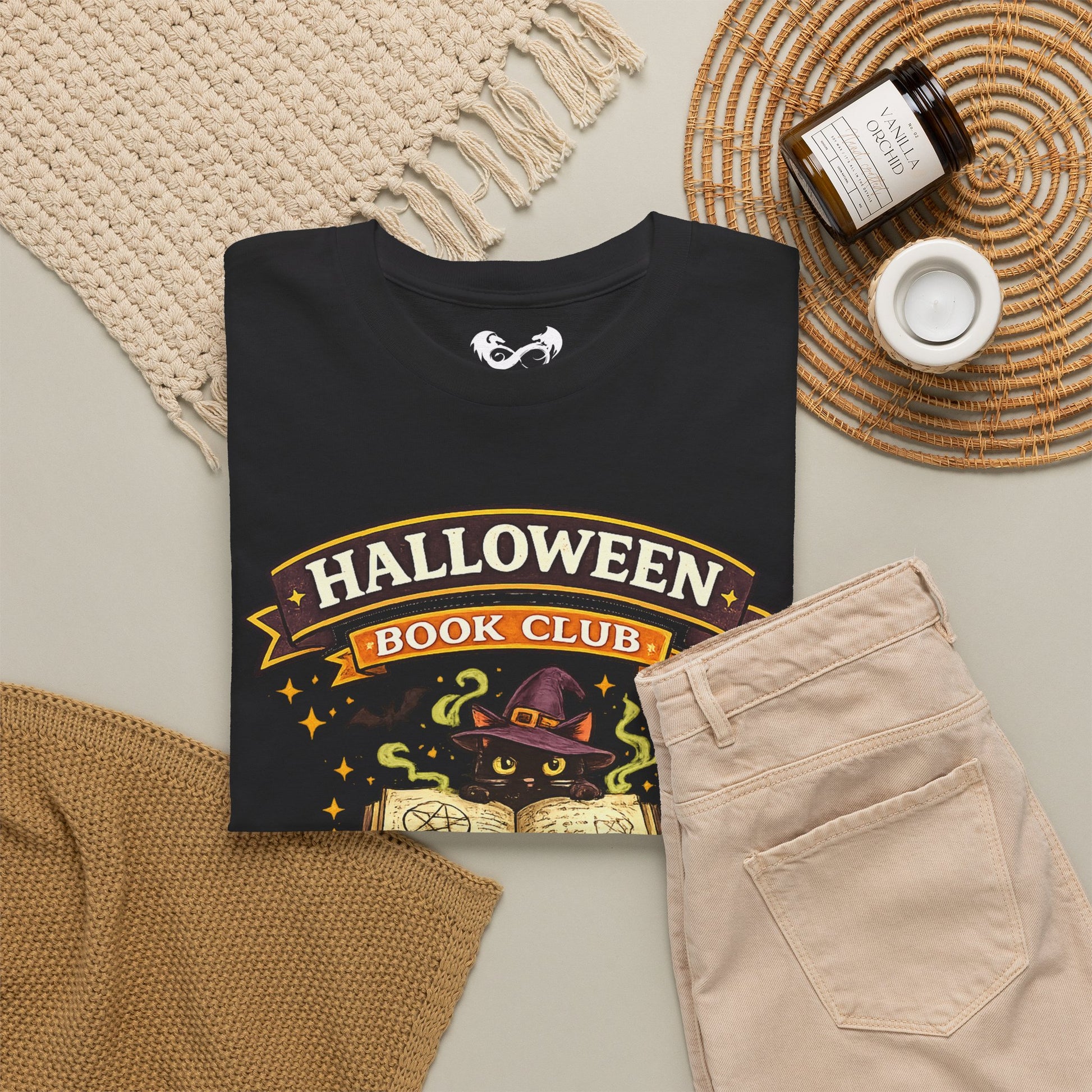 A folded black graphic T-shirt is styled in a flat lay with beige pants, a woven blanket, a small candle jar, and a cup on a neutral surface. The shirt graphic reads 'HALLOWEEN BOOK CLUB' with a witch cat and open book illustration.