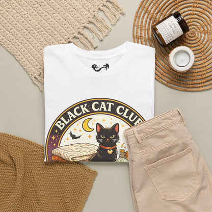 A folded white graphic T-shirt is arranged in a styled flat lay with beige pants, a woven blanket, a candle jar, and a small cup on a neutral surface. The shirt graphic shows a black cat inside a circular design with the text 'BLACK CAT CLUB' and a crescent moon.